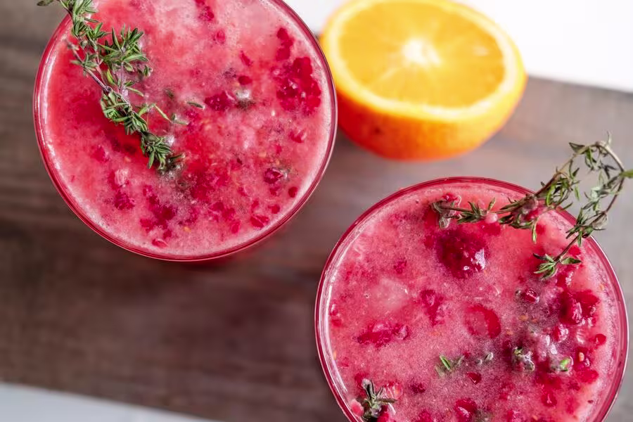 Two glasses of a red fruit beverage garnished with sprigs of thyme, with a halved orange beside them on a wooden surface.