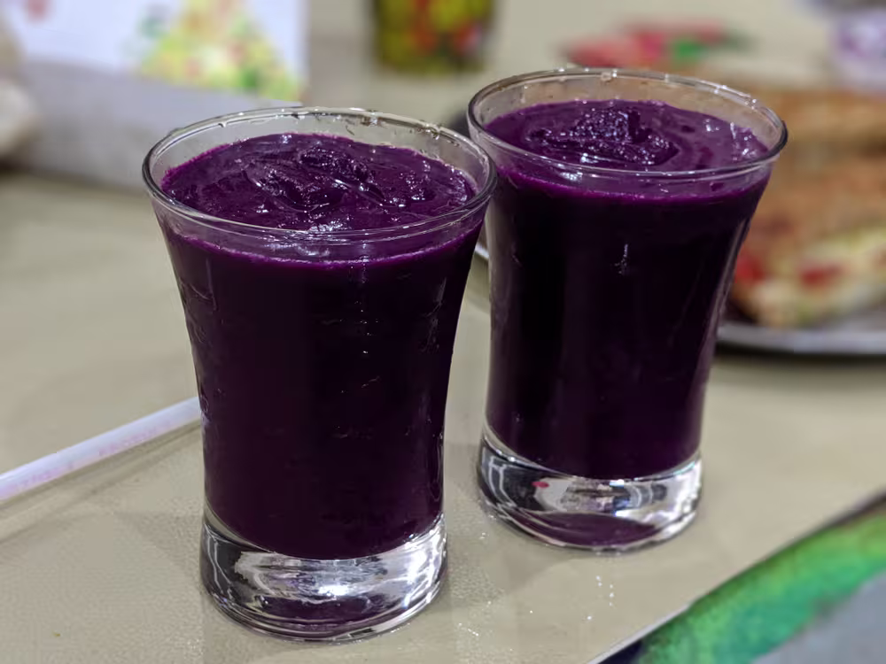 Two clear glasses filled with thick, dark purple smoothie are placed on a table with food items visible in the background.