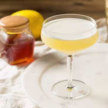 A yellow cocktail in a coupe glass sits on a tray with a metal jigger, a lemon half, a cocktail shaker, and a jar of honey in the background.