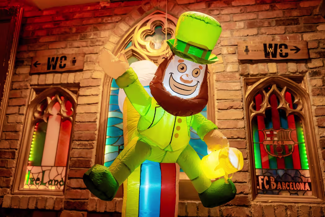 La foto muestra un evento interior concurrido con muchas personas vestidas de verde, una celebración relacionada con el Día de San Patricio. Hay decoraciones verdes y blancas, incluyendo banderas y guirnaldas, colgadas por el techo. 