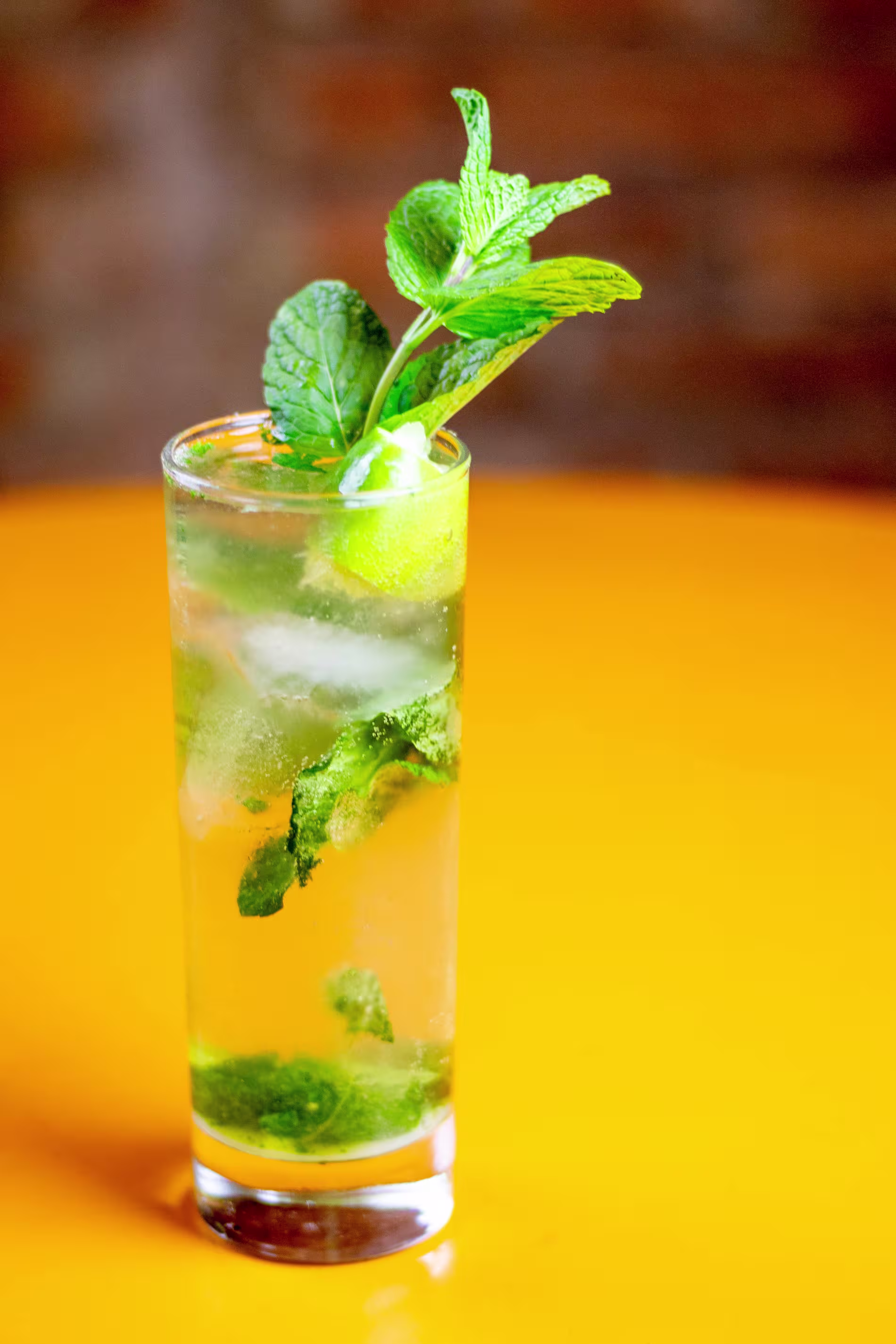 A tall glass of mojito with ice, lime wedges, and fresh mint leaves, set on an orange surface with a blurred background.