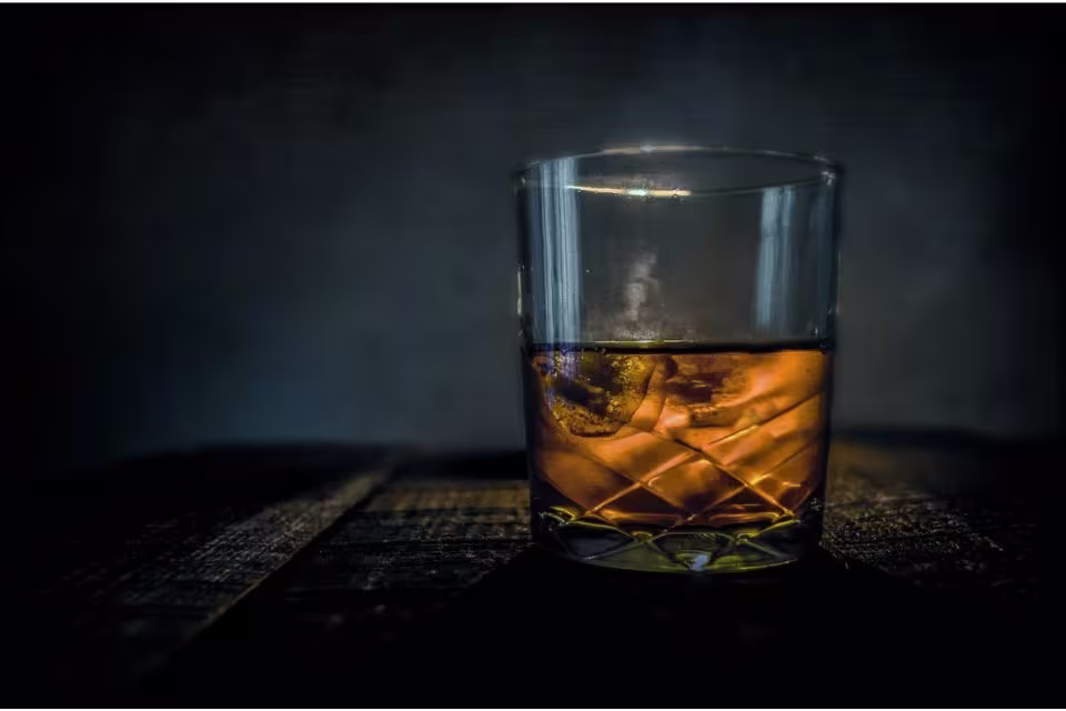 A glass of whiskey with ice sits on a dark wooden surface against a dimly lit background.