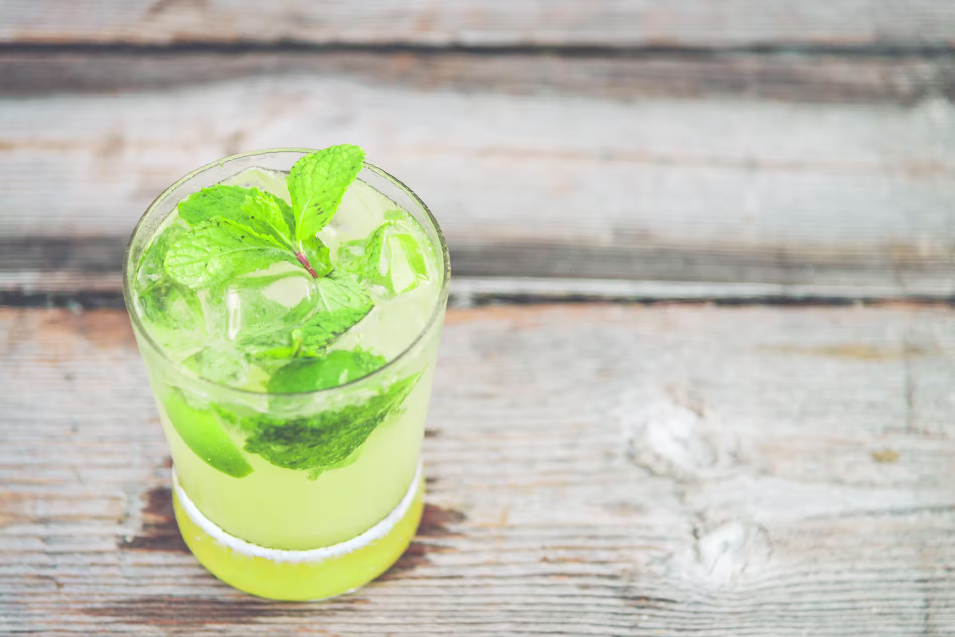A glass of iced lime drink garnished with fresh mint leaves sits on a weathered wooden surface.