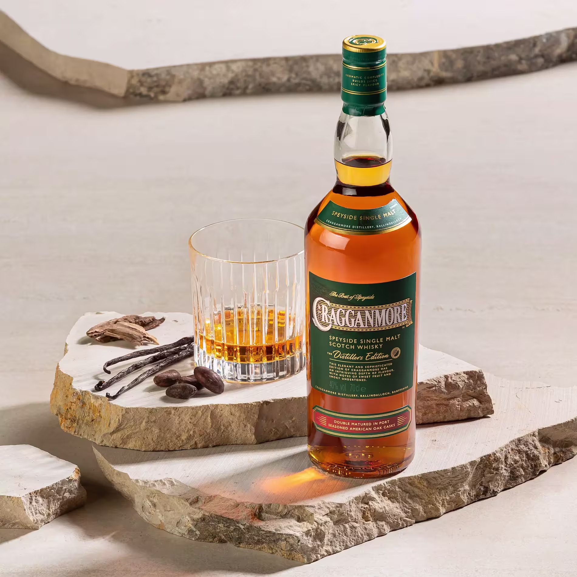 A bottle of Cragganmore single malt Scotch whisky stands next to a glass of whisky and a few vanilla pods and cocoa beans on stone slabs.