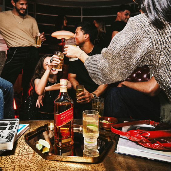 La imagen muestra una botella de whisky Johnnie Walker Red Label sobre una mesa y, alrededor, un grupo de personas brindando con vasos rellenos con cócteles preparados con dicho destilado.
