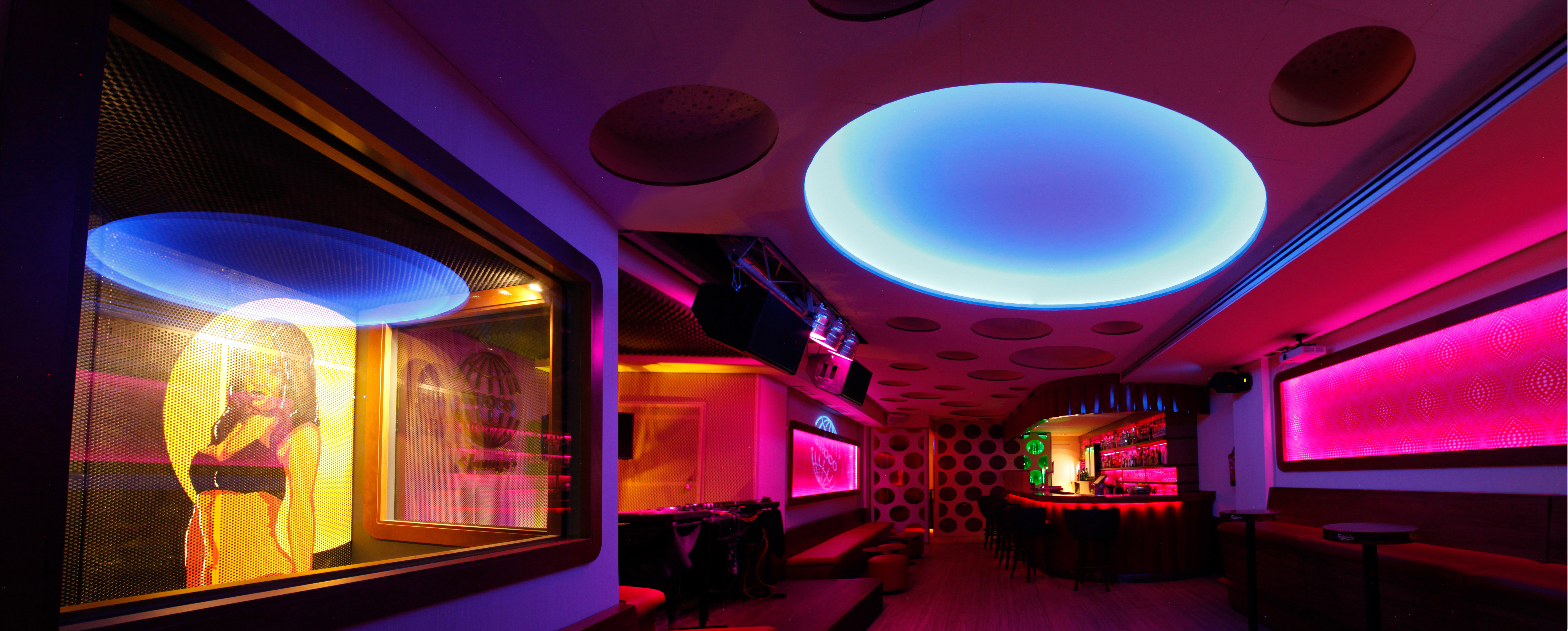La imagen muestra el interior de la Sala Sirocco, con una decoración moderna y elegante. Hay una barra iluminada con luces LED, varias botellas en estantes detrás de la barra, y taburetes altos para los clientes. Las paredes luces colgantes