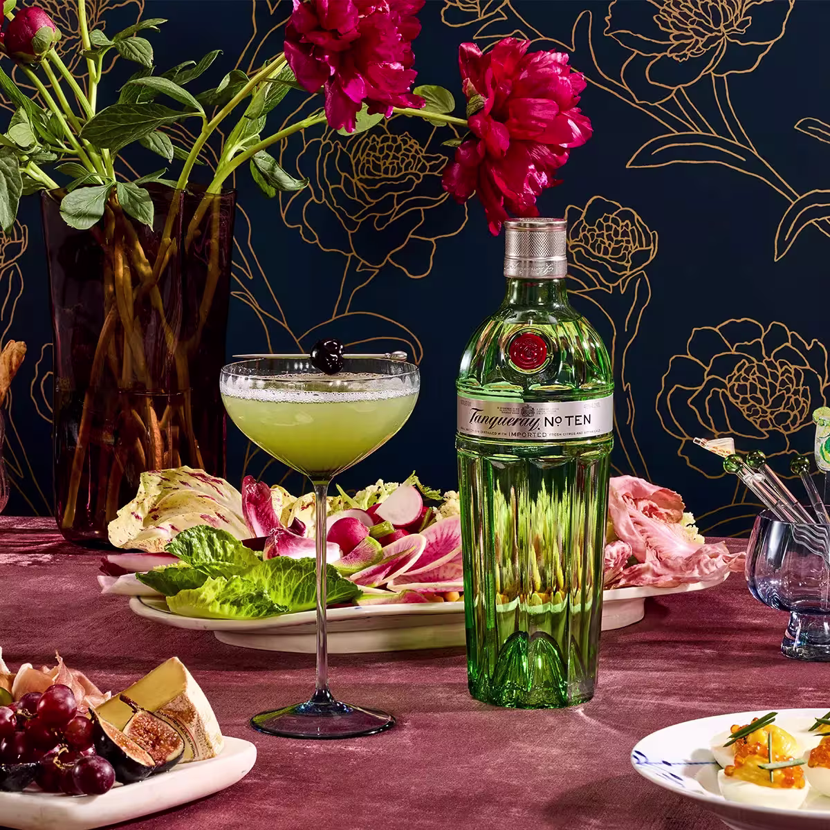 A bottle of Tanqueray No. Ten gin next to a cocktail glass with a cherry garnish, surrounded by plates of fresh vegetables, fruit, and flowers on a patterned table.