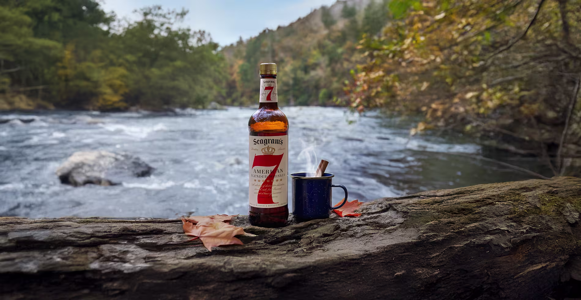 A bottle of Seagrams 7 Crown whiskey, a blue enamel mug, and autumn leaves rest on a log beside a flowing river in a forested area.