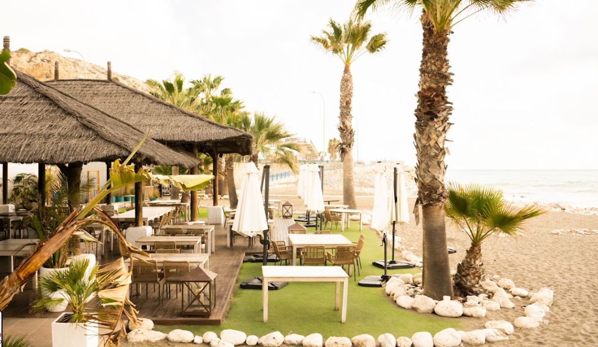Una mesa en el restaurante Candado beach puesta cerca de una ventana abierta que ofrece una vista de la playa y el océano. El lugar parece estar en una ubicación cálida y soleada, dado la presencia de palmeras y la luz brillante del sol. 