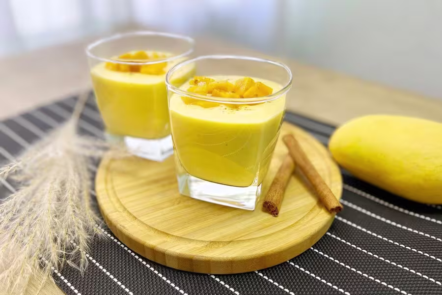 Two glasses of mango mousse topped with diced mango sit on a wooden tray beside a whole mango and two cinnamon sticks.
