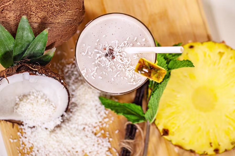 A glass of creamy smoothie topped with shredded coconut, surrounded by coconut halves, pineapple slice, mint leaves, and a wooden board.