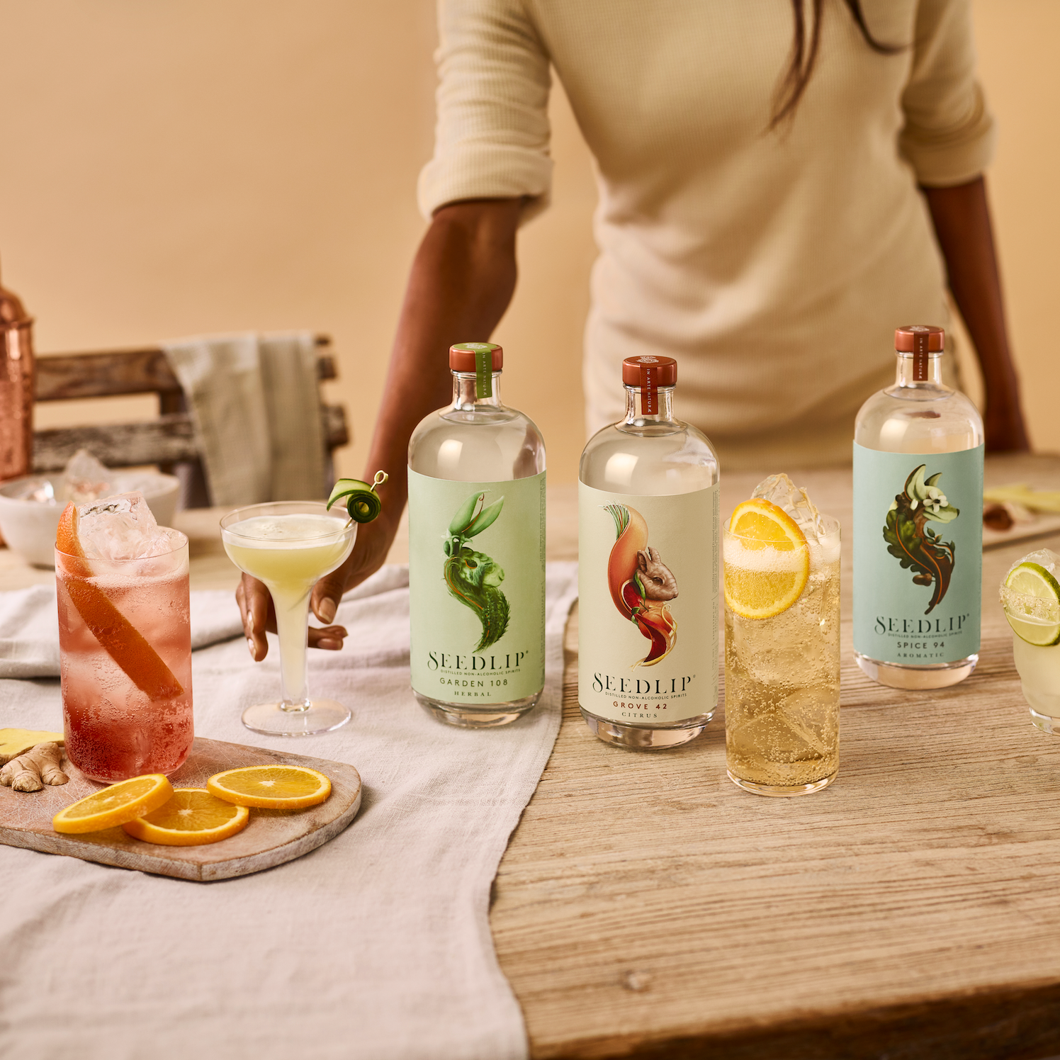 Seedlip bottles and cocktails on a wooden table with a tablecloth and flowers.