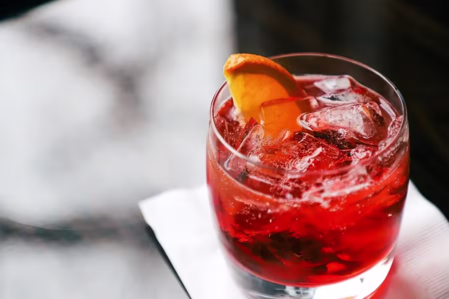 A glass of red cocktail with ice cubes and an orange wedge garnish sits on a white napkin.