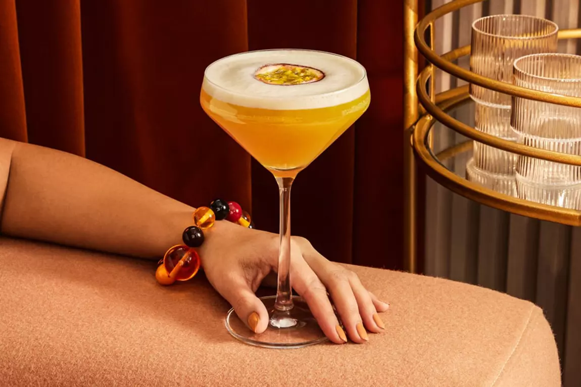 A hand rests on a peach-colored surface, holding a coupe glass filled with a yellow cocktail topped with foam and a halved passion fruit. Stacked glasses and a gold bar cart are in the background.