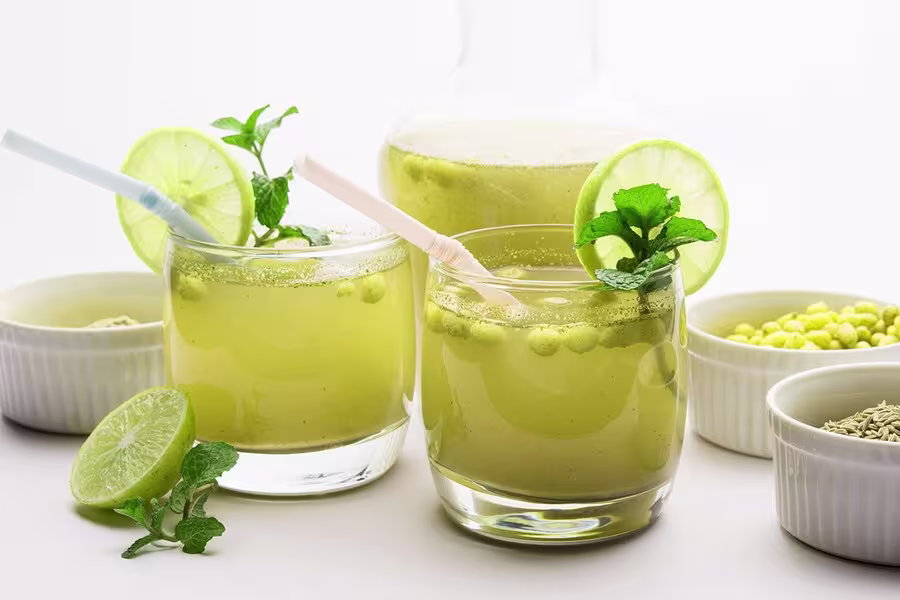 Two glasses of green lemonade with mint and lime slices, each with a straw, surrounded by bowls of ingredients on a white surface.