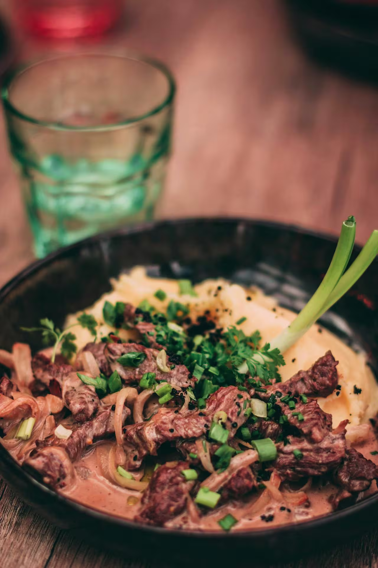 A black bowl contains beef stroganoff served over mashed potatoes, garnished with chopped chives and green onions, with a glass of water in the background.