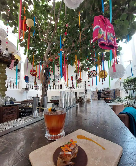 En la imagen se ve la barra del Astrid, con un cóctel y una pequeño plato de comida. Es en el exterior y los arboles están decorados con cosas autóctonas.