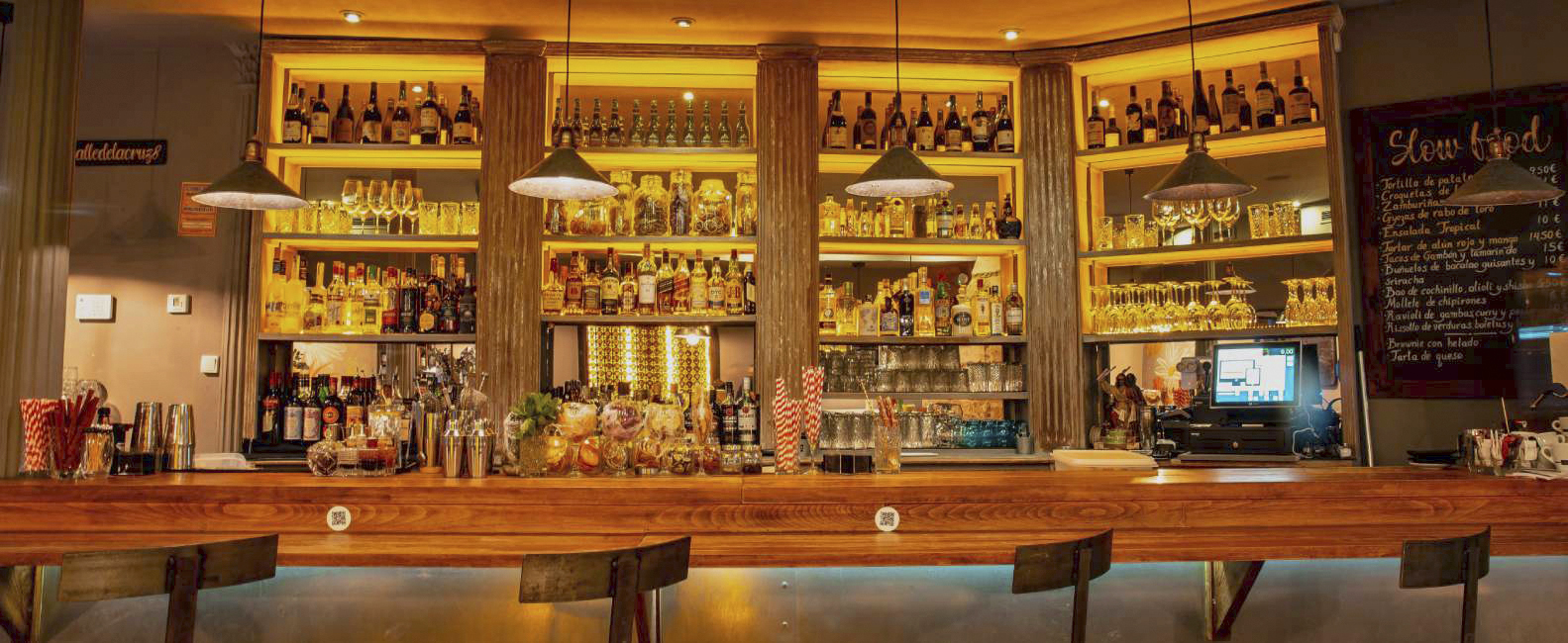 La imagen muestra la barra de Bandida, decorada con estantes llenos de botellas de licor y copas de vidrio cuidadosamente dispuestas. La iluminación cálida y las columnas de estilo clásico crean una atmósfera acogedora y elegante.