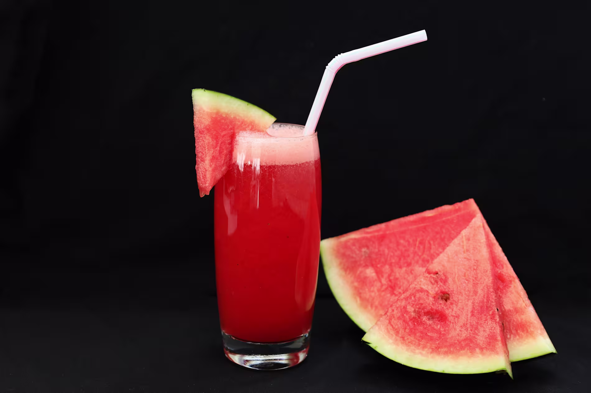 A glass of watermelon juice with a straw and a watermelon slice garnish, next to three watermelon wedges on a black background.