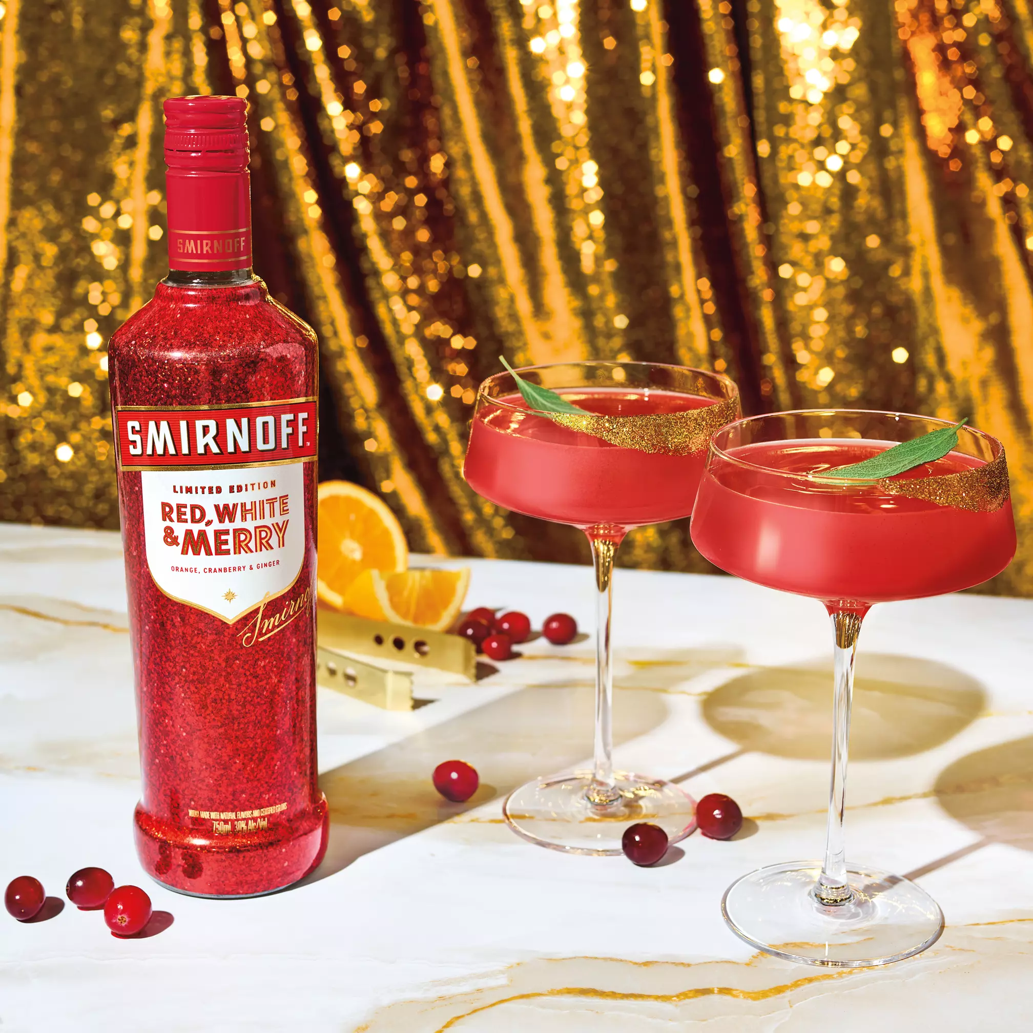 A bottle of Smirnoff Red, White & Merry vodka sits beside two red cocktails in coupe glasses, with cranberries, orange slices, and gold sequin fabric in the background.