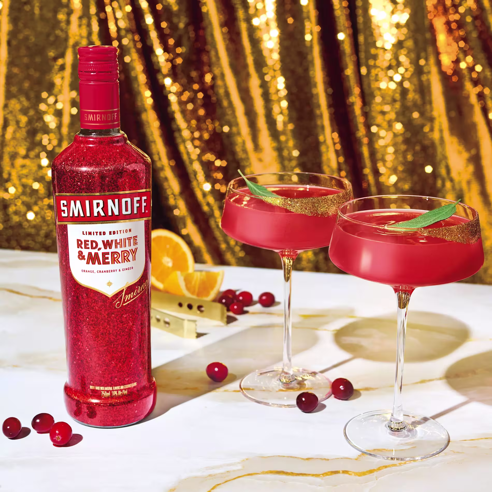 A bottle of Smirnoff Red, White & Merry vodka sits beside two red cocktails in coupe glasses, with cranberries, orange slices, and gold sequin fabric in the background.