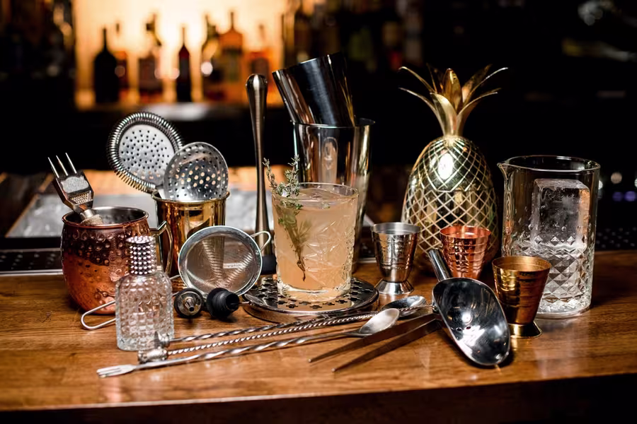 A variety of cocktail-making tools and glasses arranged on a wooden bar counter, with a mixed drink in the center and bottles blurred in the background.