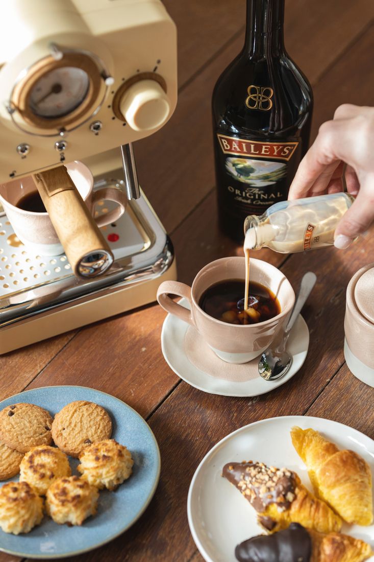 La imagen muestra una cafetera, una botella de Baileys Original Irish, dos tazas con café y dos platos con alimentos dulces en ellos.