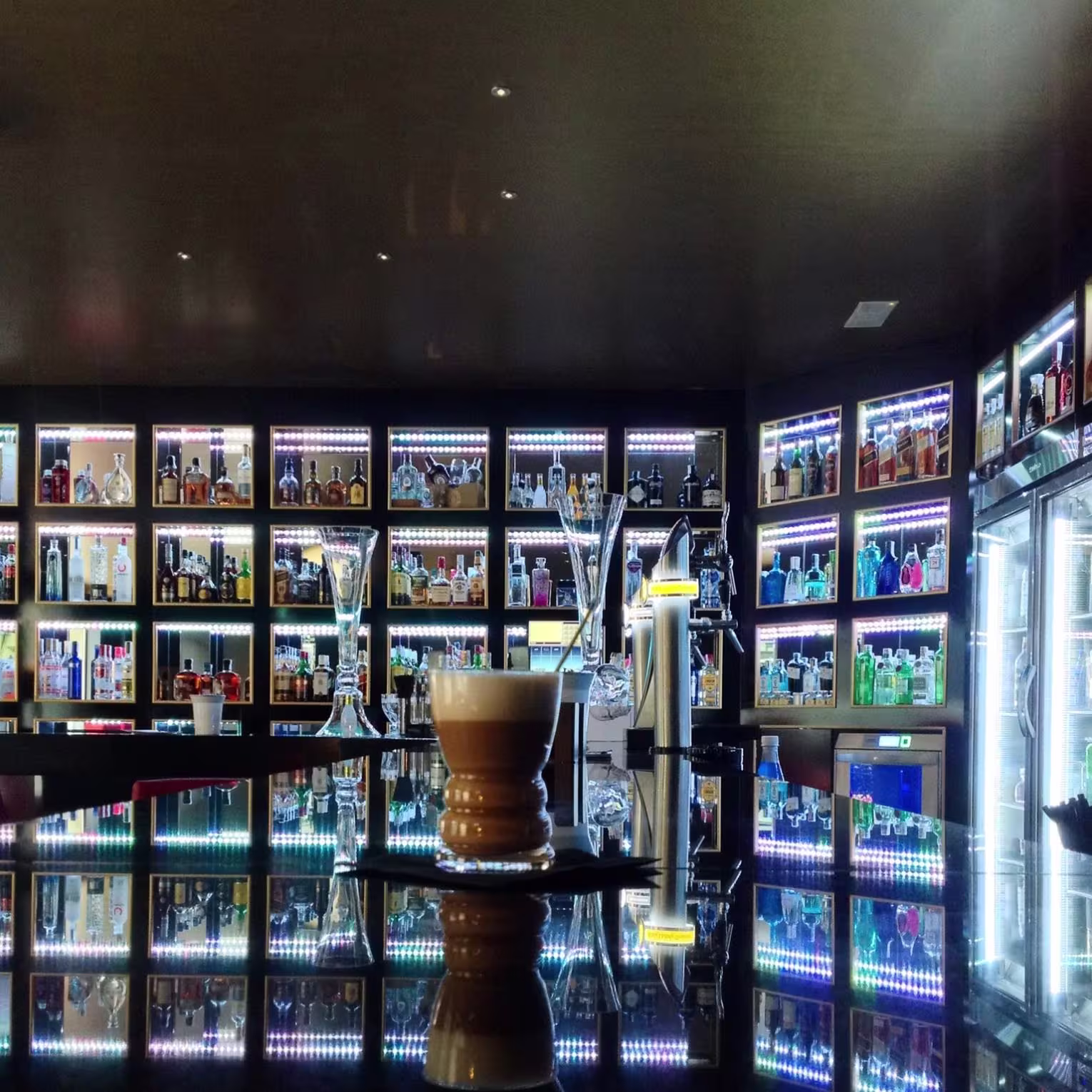 Reflejos brillantes en la barra del Majestic Premium Bar, con estanterías llenas de botellas iluminadas y una copa en primer plano sobre una superficie pulida.
