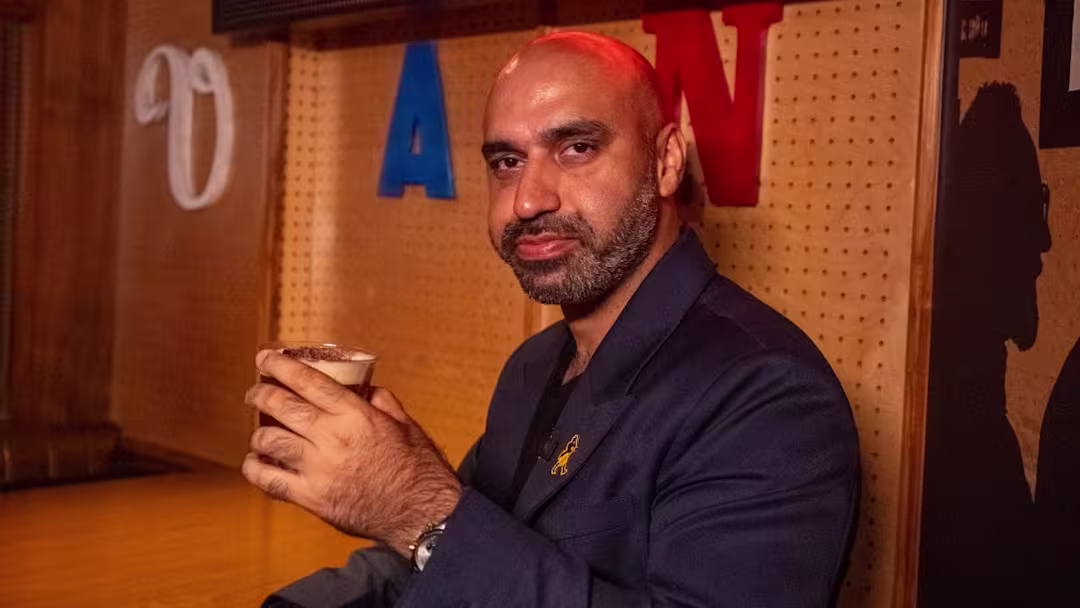 A hand holds a glass of dark liquid, likely an alcoholic beverage, over a wooden surface with blurred blue and orange lights in the background.