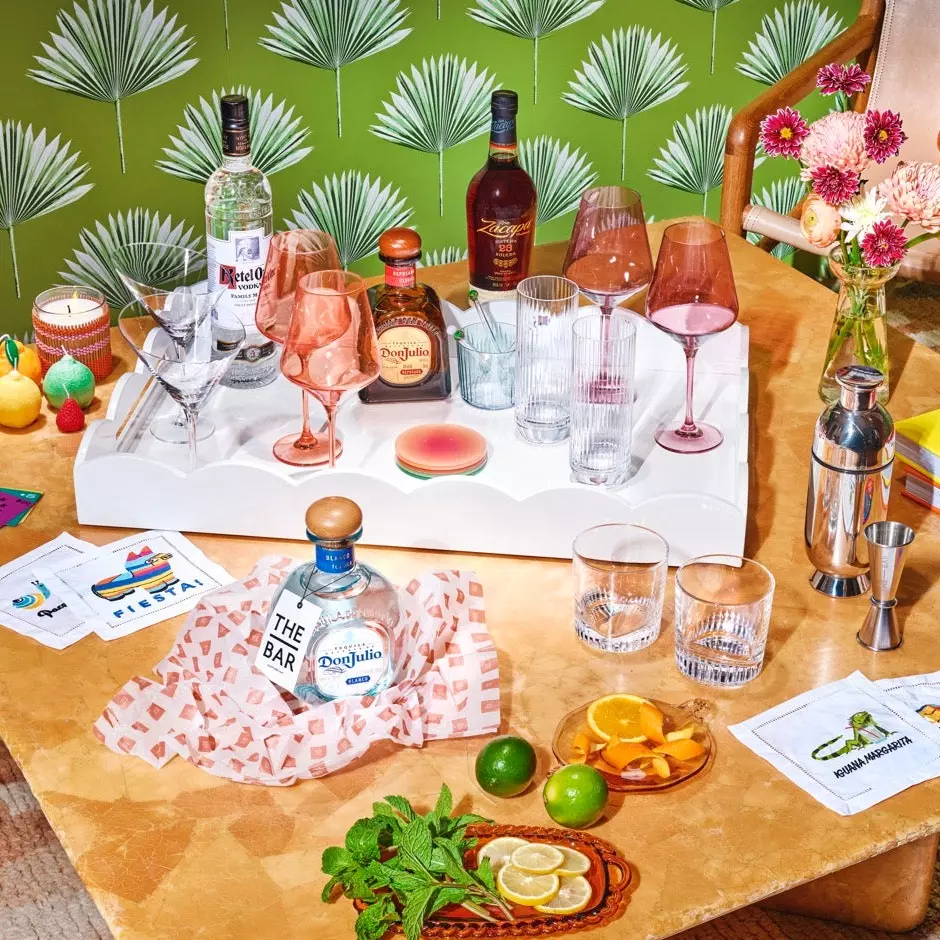 A tabletop bar setup with bottles of liquor, assorted glasses, sliced citrus fruits, mint, a cocktail shaker, napkins, and flowers against a green patterned background.