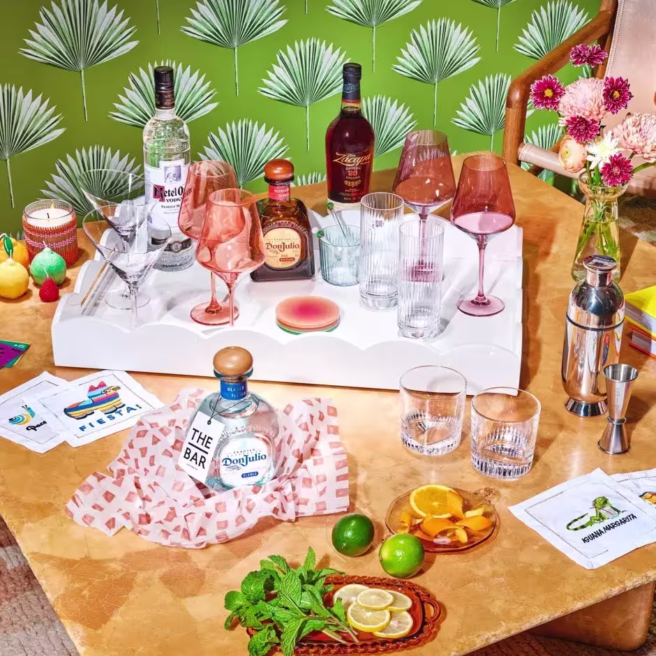 A tabletop bar setup with bottles of liquor, assorted glasses, sliced citrus fruits, mint, a cocktail shaker, napkins, and flowers against a green patterned background.