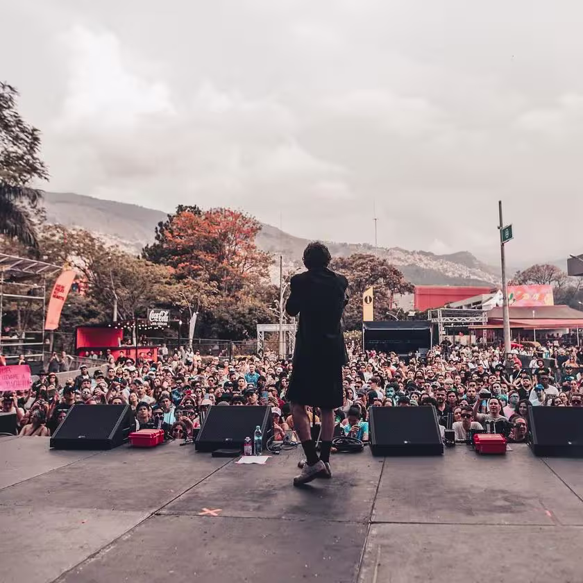 Un cantante sobre el escenario canta frente a la multitud. De fondo se ven unas montañas.