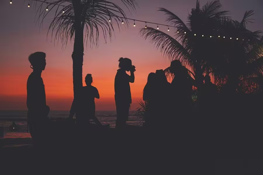 Silhouettes of people standing by palm trees at a beach during sunset, with string lights overhead and an orange sky in the background.