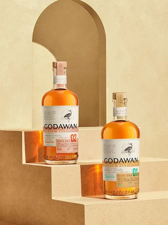 Two Godawan whisky bottles are displayed on beige stepped platforms with an arched cutout in the background, under soft lighting.