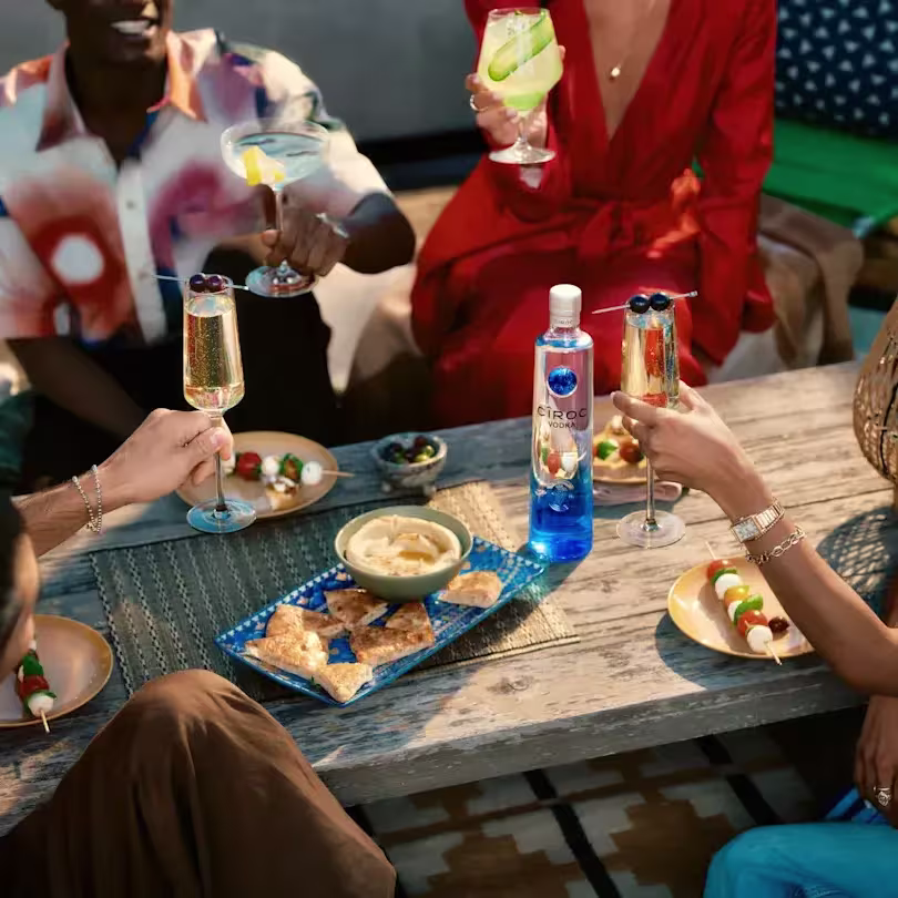 Cuatro personas brindan con cócteles alrededor de una mesa de madera con comida, que incluye hummus, patatas fritas de pita y una botella azul de vodka Ciroc.