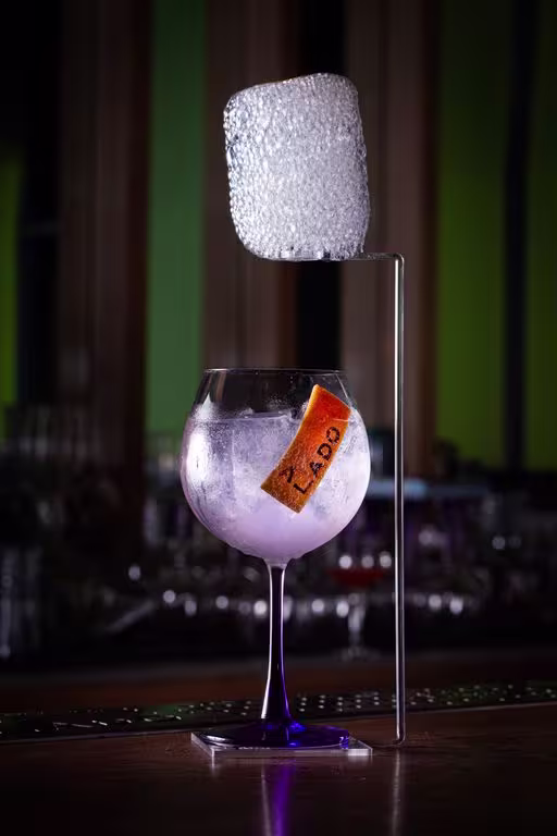 En la imagen se aprecia una copa con Gin Tanqueray London Dry, adornada con una lámina de naranja que lleva la inscripción 'Alado', sobre una barra de bebidas. En el exterior de la copa pero unido a su base, se eleva un soporte que sostiene una nube de burbujas.