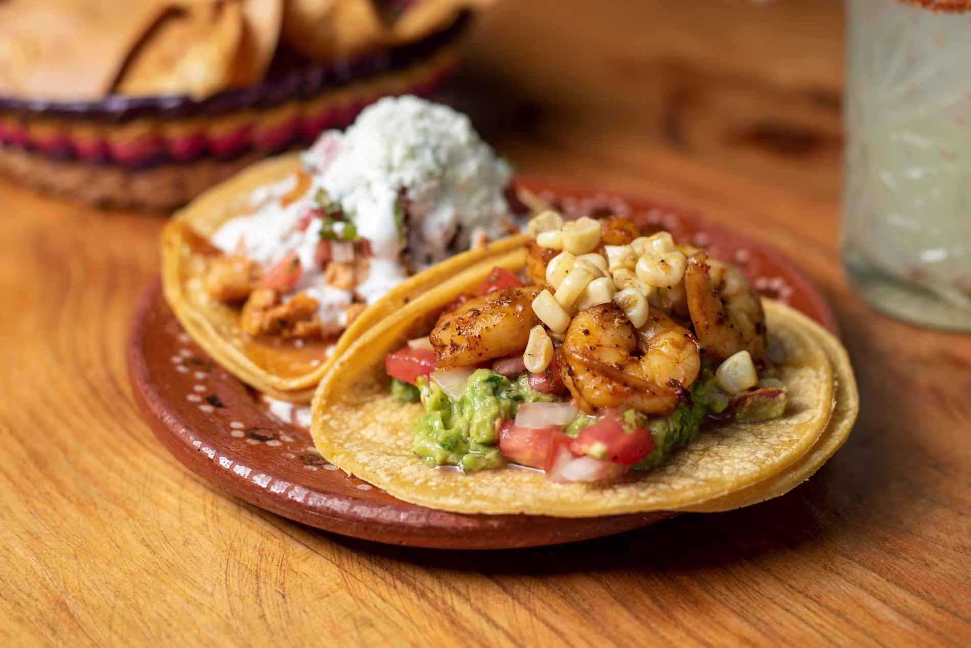 Two corn tortillas topped with shrimp, corn, guacamole, diced tomatoes, and cheese sit on a decorative plate, with chips and a drink in the background.