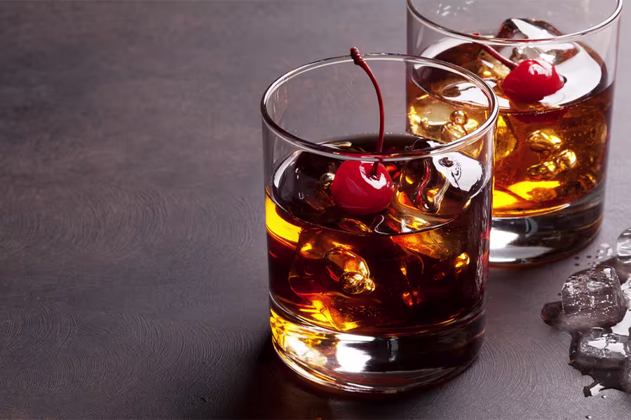 Two glasses of whiskey with ice cubes and maraschino cherries as garnish on a dark surface, with some ice pieces scattered nearby.