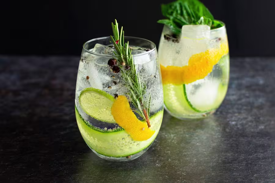 Two clear glasses of gin and tonic with ice, garnished with cucumber slices, orange peel, rosemary, basil leaves, and juniper berries, on a dark surface against a black background.