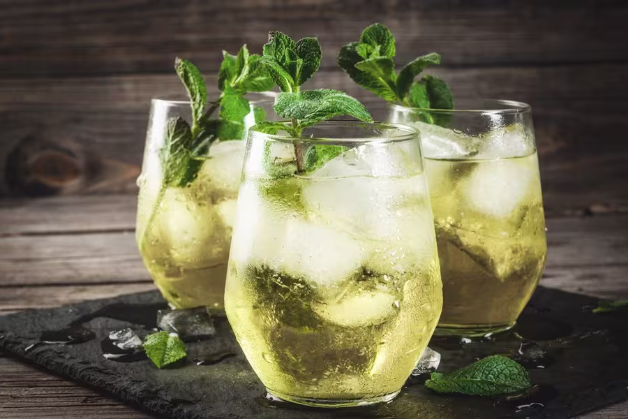 Three glasses of iced yellow drink garnished with fresh mint leaves sit on a dark slate surface with scattered ice and mint, against a wooden background.