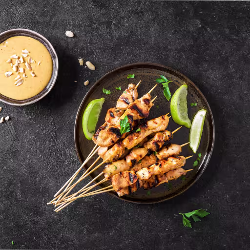Grilled chicken skewers on a dark plate with lime wedges and herbs, served next to a bowl of peanut sauce on a black textured surface.
