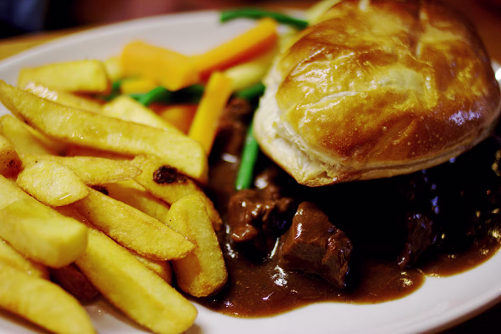 Plate with thick-cut fries, a meat pie topped with pastry, brown gravy, and a side of steamed carrots and green beans.