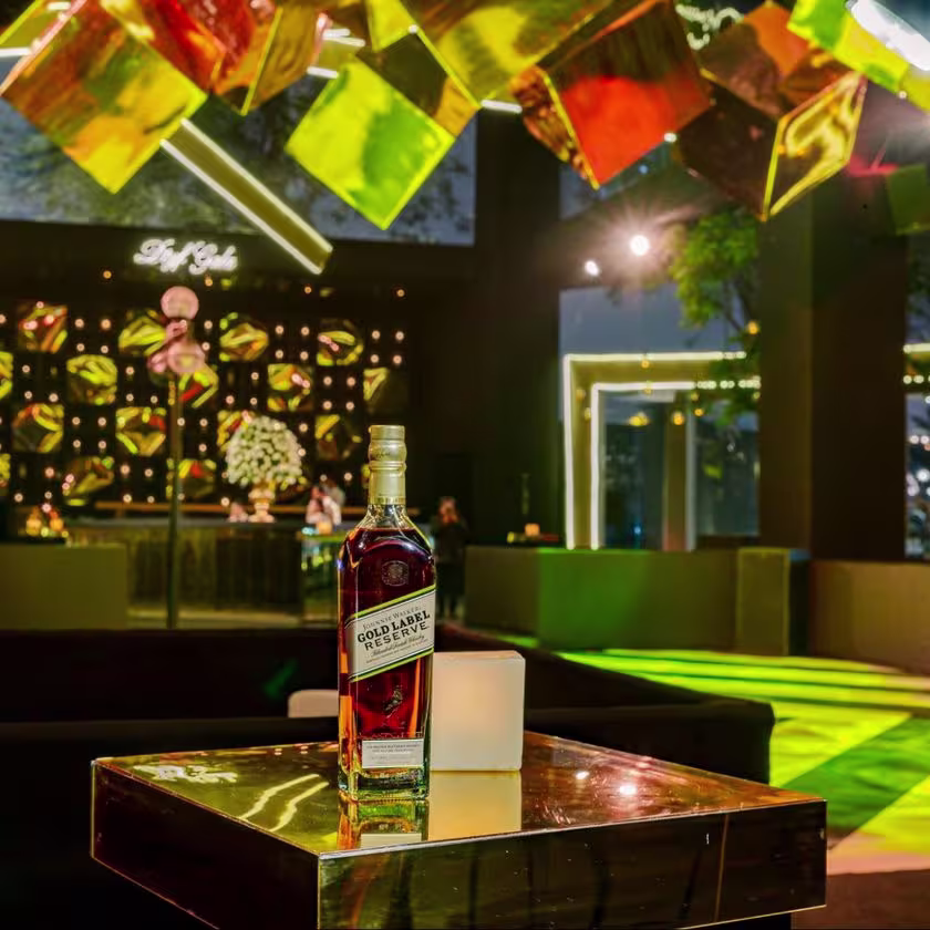 La imagen muestra la botella de whisky Johnnie Walker Gold Label Reserve sobre una mesa y, de fondo, la pista de baile.
