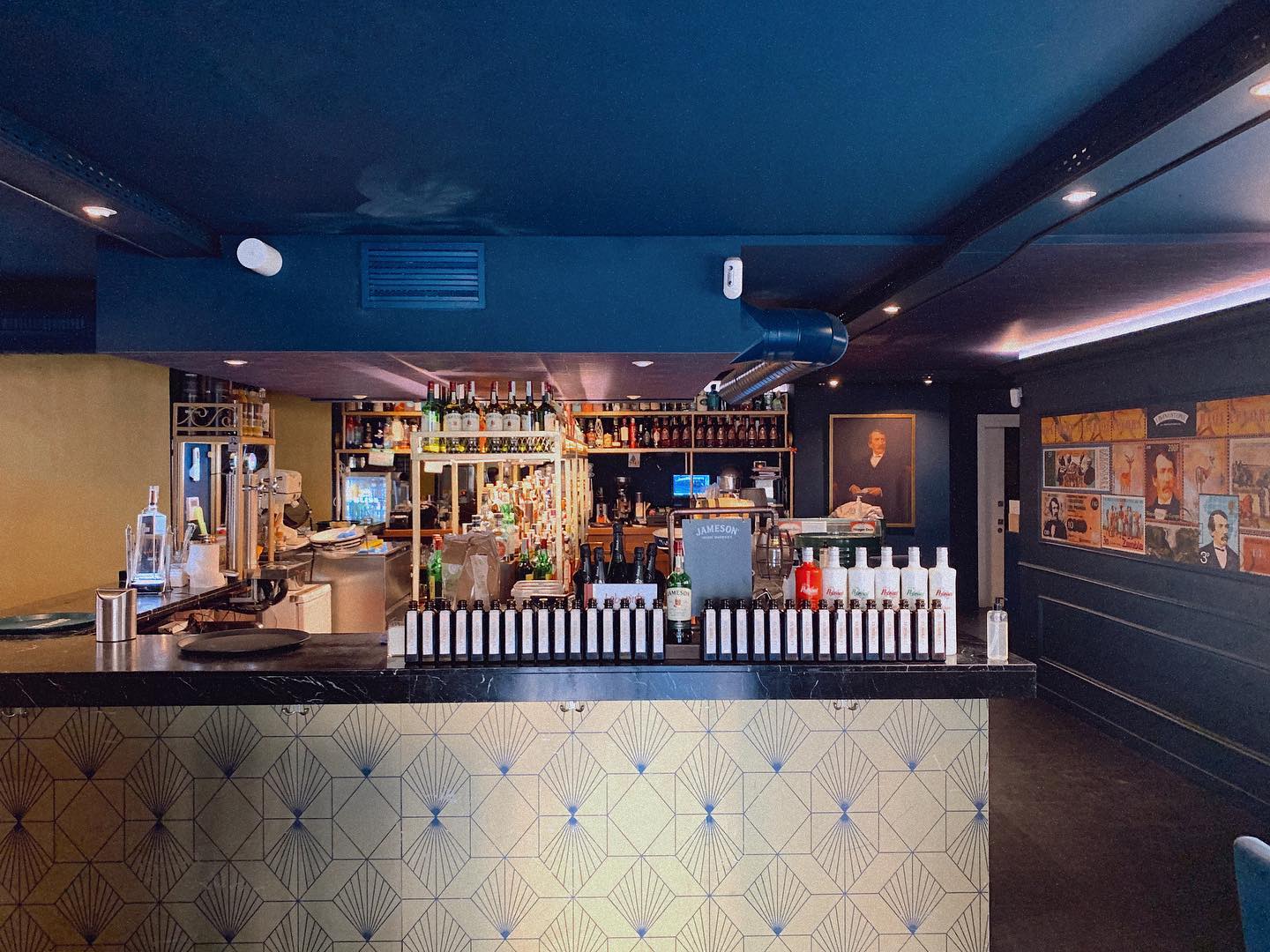 El Livingstone es un bar de ambiente íntimo y elegante, con una barra central decorada en tonos dorados y azul oscuro. Botellas de alta gama y retratos en las paredes añaden un toque vintage y acogedor.