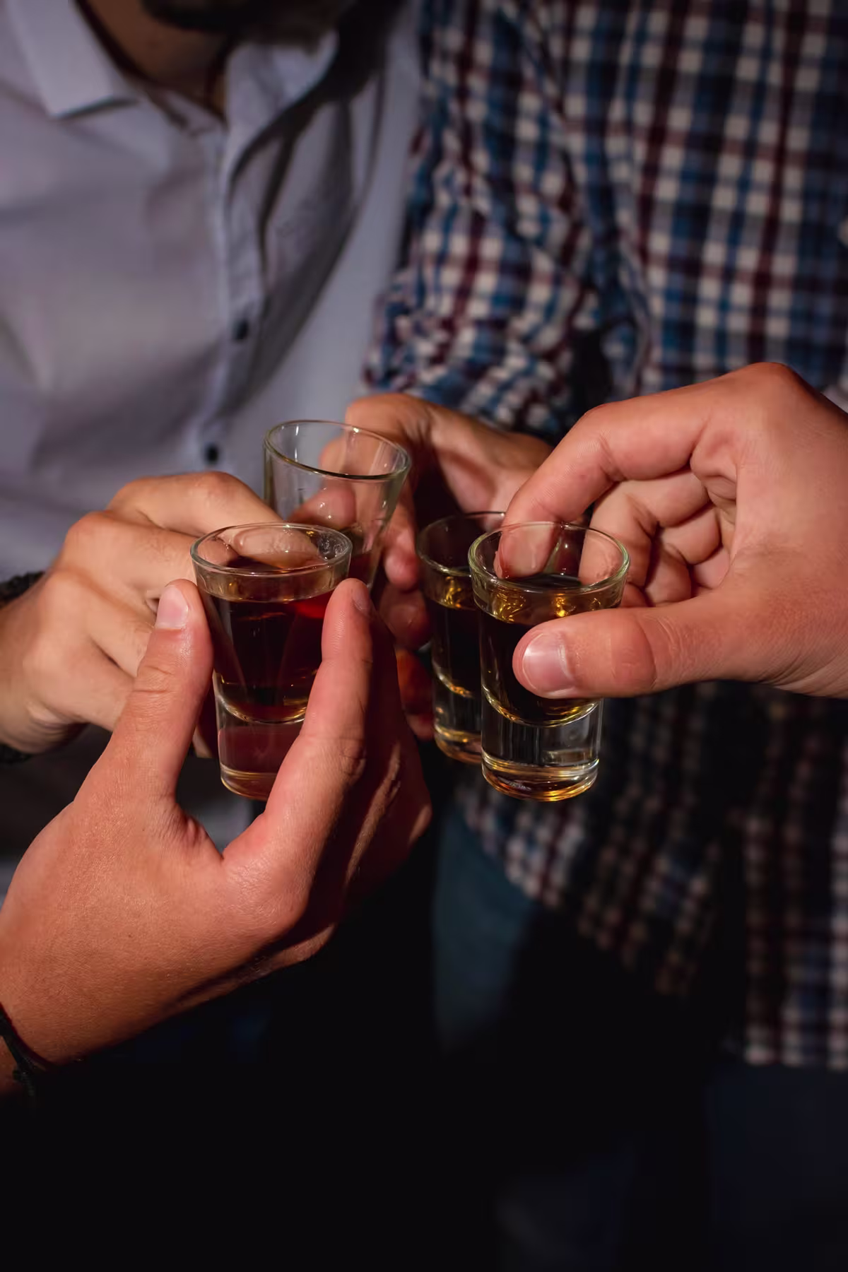 Four people clinking shot glasses filled with dark liquor, with only their hands and torsos visible.
