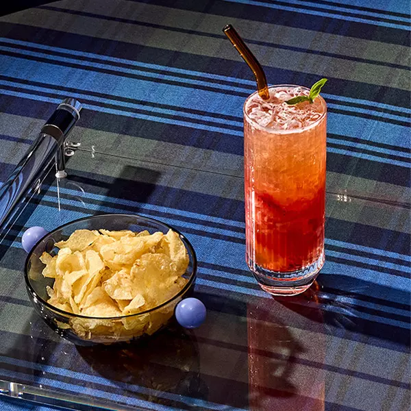 A tall glass of red, iced beverage with a straw and mint garnish sits beside a bowl of potato chips on a blue plaid tablecloth.