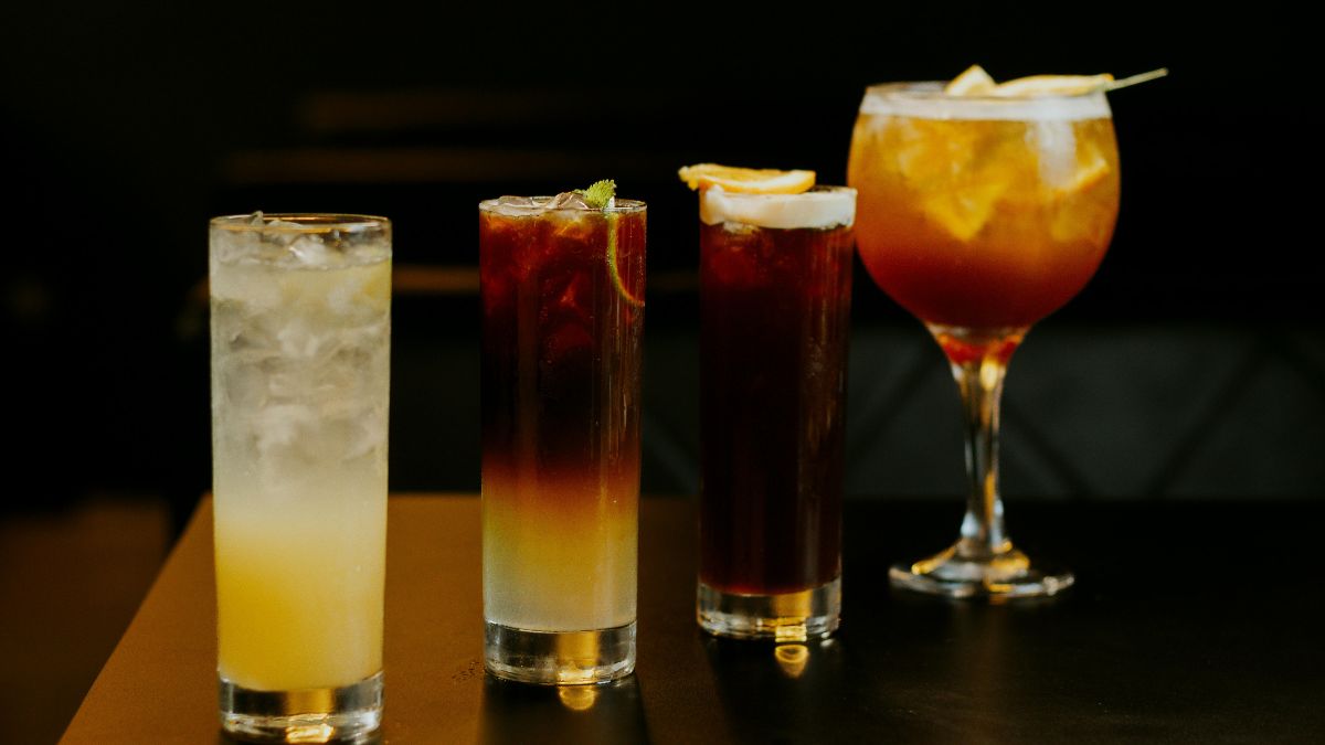 Four different cocktails in various glasses are arranged in a row on a dark surface, each garnished with citrus slices, against a dark background.
