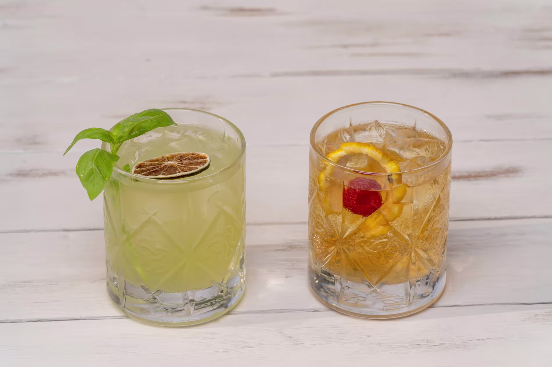 Two cocktails on a white wooden surface; the left drink is light green with a basil leaf and dried lime garnish, the right drink is amber with orange and cherry garnish.