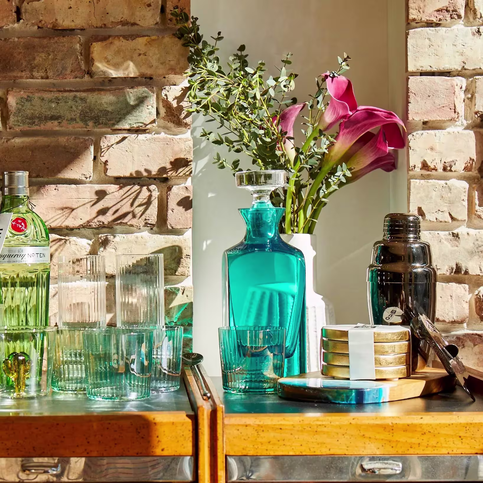 A bar cart with gin, glassware, a blue decanter, cocktail shaker, stacked coasters, and a vase with pink flowers and greenery against a brick wall.
