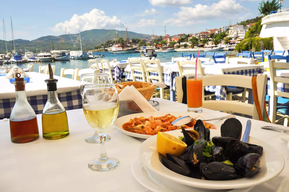 A table set with seafood dishes, wine, and juice overlooks a marina with boats and hills in the background on a sunny day.